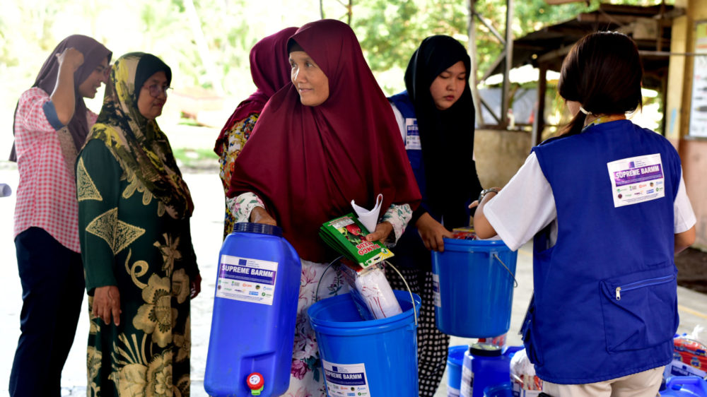 A woman wearing a maroon burka carries two blue plastic containers of water