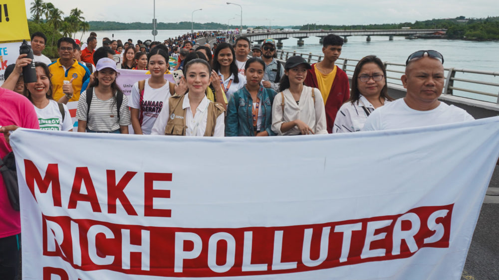 Hundreds of people are walking across a long bridge. The people in front are carrying a banner that says "Make rich polluters pay now" in red font.