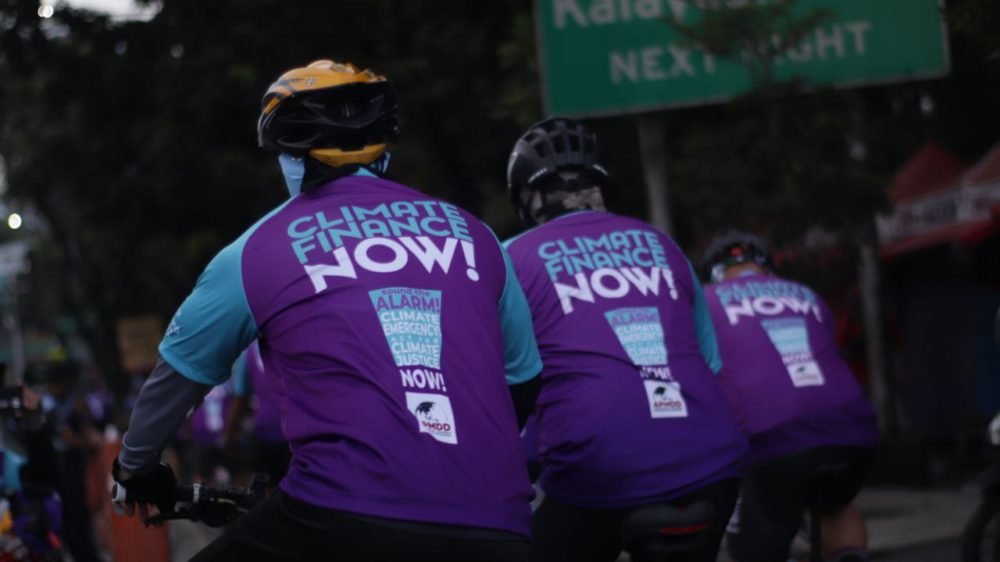 Three cyclists wearing purple jerseys with the words "Climate Finance Now!" on their backs. There is a street sign that says "Kalayaan Avenue next right" on the background.