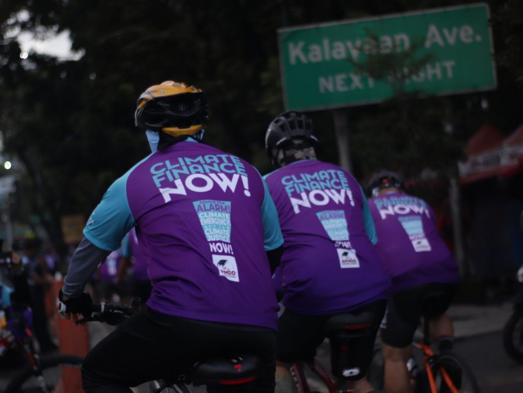 Three cyclists wearing purple jerseys with the words "Climate Finance Now!" on their backs. There is a street sign that says "Kalayaan Avenue next right" on the background.