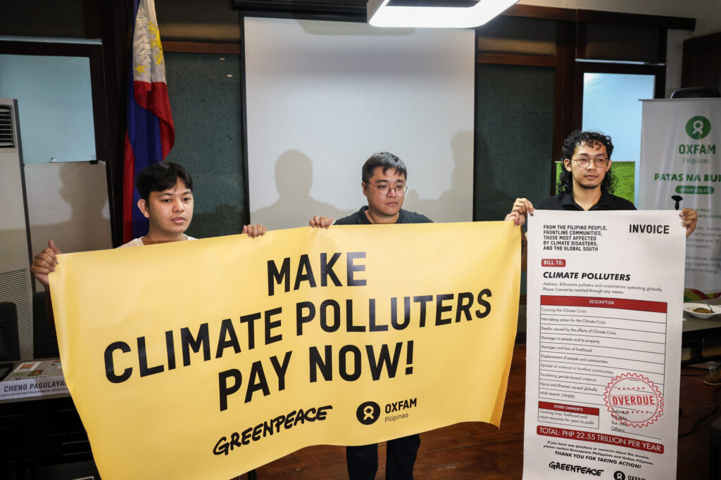 Two persons in front of a conference room holding banners that say "Make Climate Polluters Pay Now!" with the Oxfam Pilipinas and Greenpeace logo below. To the right, another person holds an enlarged symbolic invoice "charged" to climate polluters.